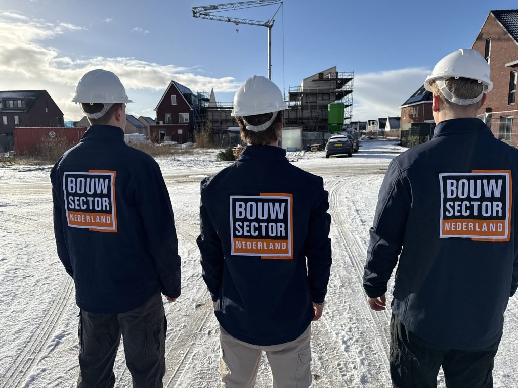 Drie vakmensen met witte bouwhelm kijken naar woningen in aanbouw met steigers op de achtergrond